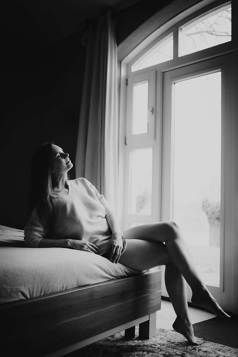 Zwart-wit boudoirportret bij een groot raam, zacht invallend daglicht.